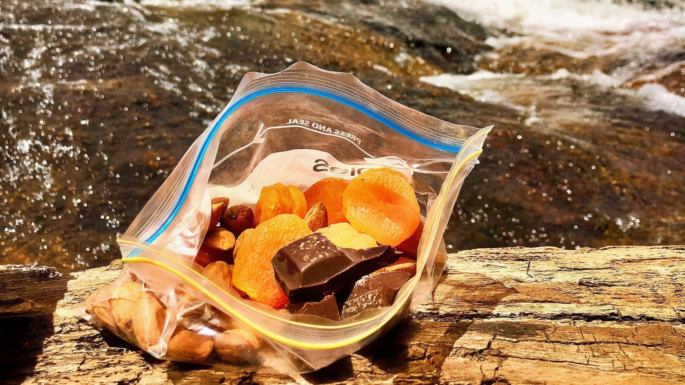 Resealable bag of trail mix with nuts, dried fruit and dark chocolate beside a stream